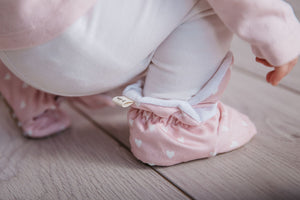 Baby girl playing wearing a pair of pink stay-on baby booties featuring a white heart print on cotton fabric, with non-slip soles and adjustable three snap closure. Made from organic cotton.