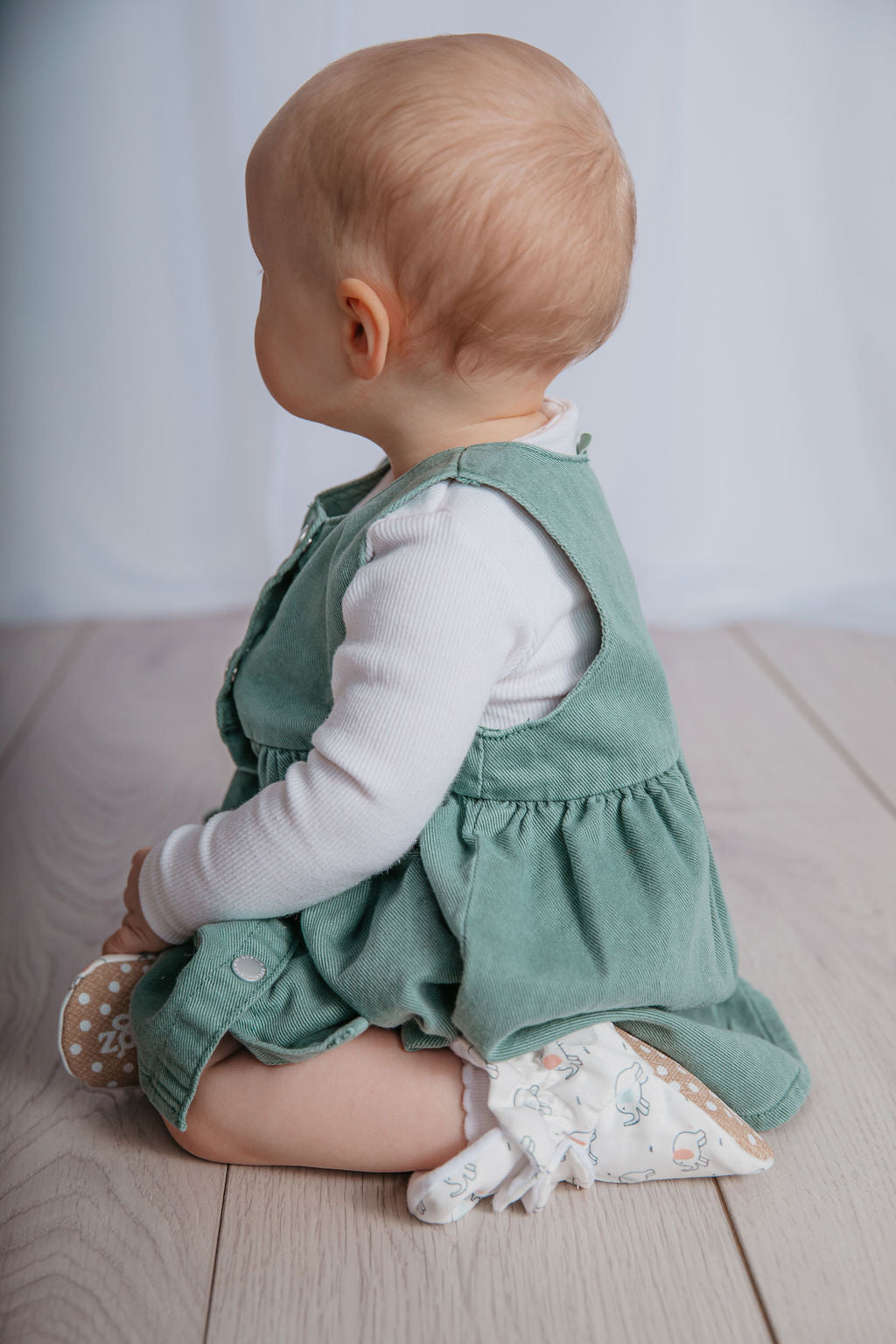 Baby girl sitting wearing a pair of stay-on baby booties featuring an elephant print on white cotton fabric, with non-slip soles and adjustable three snap closure. Made from organic cotton.