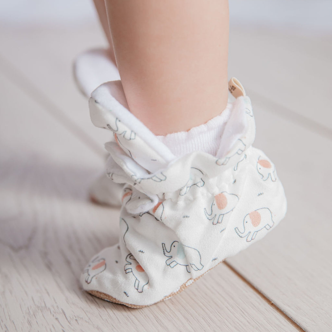 Baby girl on tip toes wearing a pair of stay-on baby booties featuring an elephant print on white cotton fabric, with non-slip soles and adjustable three snap closure. Made from organic cotton.