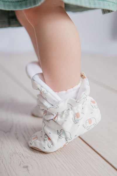Baby girl on tip toes wearing a pair of stay-on baby booties featuring an elephant print on white cotton fabric, with non-slip soles and adjustable three snap closure. Made from organic cotton.