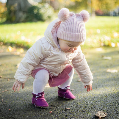 Child in a pink coat and hat wearing Snugabugz plum water-resistant booties playing outdoors on a grassy area.