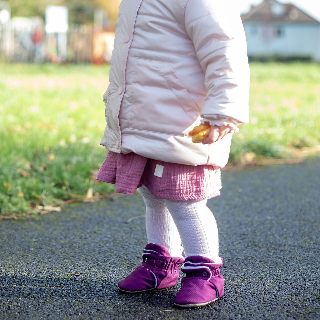 Child wearing a pink coat, pink dress, and purple water-resistant stay on baby booties standing on a path with grass and a playground in the background.