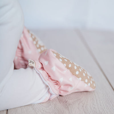 Baby girl crawling wearing a pair of Snugabugz pink baby booties that stay on featuring a white heart print on organic cotton fabric, with grip soles and adjustable snap closure. Made from organic cotton. pre-walker baby shoes.