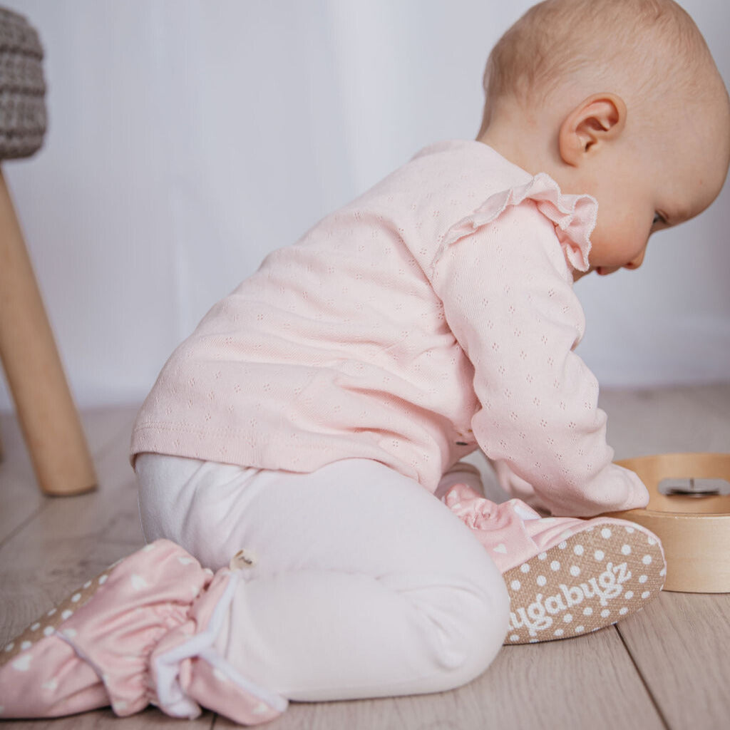 Baby girl playing wearing a pair of pink Snugabugz stay-on infant baby booties that don't fall off featuring a white heart print on cotton fabric, with non-slip soles and adjustable snap closure. Barefoot baby shoes made from organic cotton.